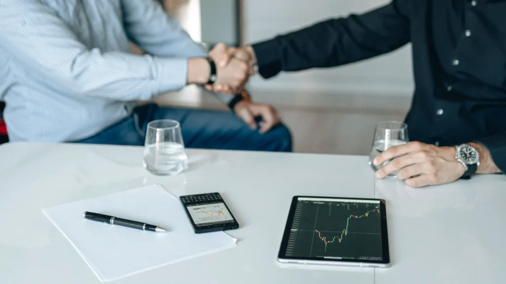 Two people shaking hands across a table with fund performance charts on a tablet